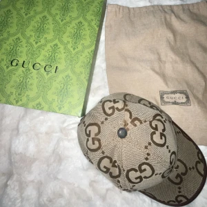 Beige keps från Gucci med GG-logga - Säljer en beige keps från Gucci med ikoniskt GG-monogram i brunt över hela kepsen. Kepsen har justerbar rem i brunt skinn baktill och är tillverkad i bomullsmix. Kommer med originalbox och dustbag. Perfekt för dig som vill sticka ut med lyxig streetstyle.