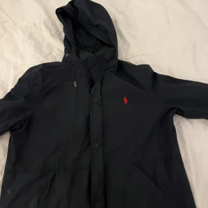 Svart vindjacka från Polo Ralph Lauren - Mörkblå vindjacka från Polo Ralph Lauren med huva och röd broderad logga på bröstet. Jackan har dragkedja framtill, bröstficka med zip och två sidofickor. Perfekt för blåsiga dagar och har en clean, sportig look.