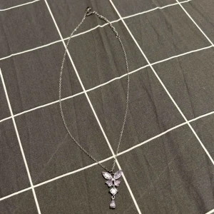 Silverhalsband med lila fjärilshänge - Snyggt halsband i silverfärgad med ett hänge som liknar en fjäril, dekorerat med lila stenar i olika former. (Inte äkta silver)