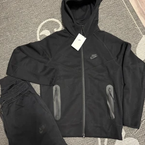 Nike tech svart - Svarta mjukisbyxor från Nike med stilren design och diskret logga på benet. Byxorna har elastisk midja med snörning, ribbade muddar vid benslut och en cool dragkedja längs sidan. Perfekta för chill eller träning, tillverkade i mjukt bomullsmaterial.