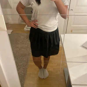 Svart volangkjol med smockmidja - Supersöt svart kortkjol med bred smock i midjan och volangig nederdel. Kjolen har en luftig och lätt form som ger en skön känsla och snygg siluett. Perfekt att styla med en enkel t-shirt eller en cool jacka.