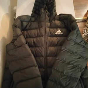 Svart pufferjacka från Adidas, L - Svart pufferjacka från Adidas med huva och dragkedja framtill. Jackan har det klassiska Adidas-logot på bröstet och är quiltad för extra värme. Perfekt för kyliga dagar. Kan behövas tvättas.