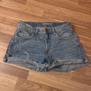 Blå denimshorts med fransig kant - Säljer ett par klassiska blå jeansshorts från Denim i storlek 38. De har fyra fickor, uppvikta och fransiga kanter för en chill vibe. Perfekta för varma dagar och har en normal passform med knapp och dragkedja framtill.