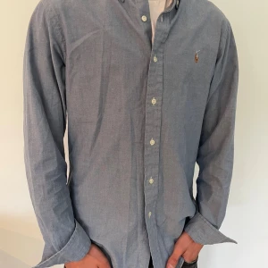 Blå skjorta från Ralph Lauren - Klassisk blå skjorta från Ralph Lauren med button-down krage och broderad logga på bröstet. Skjortan har lång ärm, knappar framtill och är tillverkad i mjuk bomull. Perfekt för en clean och stilren look.