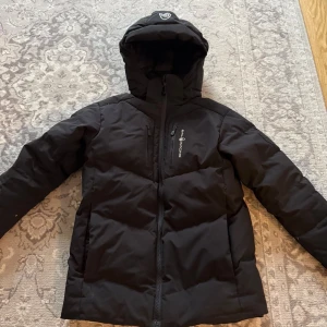 Sail Racing vinterjacka  - Svart dunjacka från Sail Racing med huva och dragkedja framtill. Jackan har en bröstficka med logga, quiltad design och är fylld med 70/30 dun/fjäder använd en hel vinter och sen så blev den för liten. 