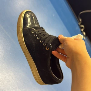 Svarta sneakers med glansig tå - Säljer ett par svarta sneakers med snörning och vit sula. Skorna har en snygg glansig tå i syntetmaterial som ger en cool kontrast mot den matta textilen. Perfekta för dig som gillar en stilren men ändå unik look.