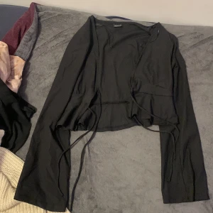 Svart omlottopp från Gina Tricot - Säljer en svart omlottopp från Gina Tricot i storlek L. Toppen har långa ärmar, ribbad struktur och knytband framtill för en snygg passform. Perfekt att styla med jeans eller kjol för en trendig look.