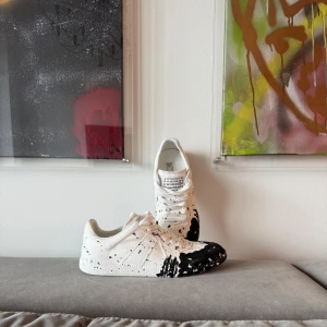 Maison Margiela skor - Maison Margiela skori i vitt med svart färgstänk-design. Klassisk låg modell med rund tå, snörning och platt sula. Skorna har en unik konstnärlig look och är tillverkade i skinn med textildetaljer. Kommer med originalkartong och dustbags.