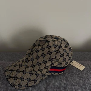 Gucci keps  - Säljer en helt ny beige keps från Gucci med klassiskt GG-mönster i mörkblått. Kepsen har en böjd skärm och en snygg detalj med blå och röd rand på sidan, samt justerbart spänne där bak. Present som aldrig kommit till användning. 