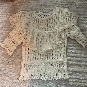 Vit stickad blus med volang från Zara - Superfin vit stickad blus från Zara med bred volang runt axlarna och ärmar i trekvarts längd. Blusen har ett öppet, virkat mönster och snygga detaljer.