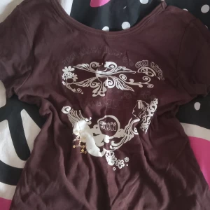 Brun t-shirt med vit print - Säljer en brun t-shirt med vit dekorativ print och blommönster framtill. T-shirten har korta ärmar och rund halsringning. Materialet känns mjukt och skönt, troligen bomull. Perfekt för dig som gillar unika tryck och avslappnad stil.