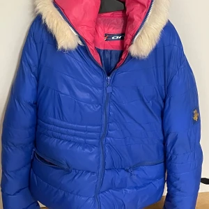 Blå pufferjacka från Carrera - Blå pufferjacka från Carrera med rosa foder och vit fuskpäls på huva och ärmslut. Jackan har dragkedja framtill, två snedställda fickor med dragkedja och broderad logga på ärmen. Perfekt för kalla dagar med sin vadderade design och snygga färgkombination.