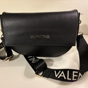 Svart axelväska från Valentino - Stilren svart axelväska från Valentino i skinn med bred, justerbar axelrem med logga. Väskan har guldfärgade metalldetaljer och flera innerfack med dragkedja. Perfekt för dig som vill ha en snygg och praktisk väska med exklusiv känsla. Det finns slitage (se bilder) inköpt på Zalando 2023.