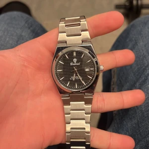 Binbond silverfärgad klocka med länk - Stilren klocka från Binbond med silverfärgad länk i metall och svart urtavla med diskret rutmönster. Klockan har datumvisning och klassisk rund form. Perfekt accessoar för dig som gillar en clean och modern look.