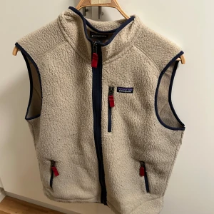 Beige fleeceväst från Patagonia L - Mysig beige fleeceväst från Patagonia med marinblå detaljer och röda dragkedjor. Västen har hög krage, tre fickor med dragkedja och klassisk Patagonia-logga på bröstet. Perfekt för lager-på-lager och outdoor-vibe. Använt flertal gånger, men ändå bra skick. Vid frågor skriv till mig!