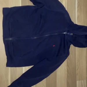 Marinblå hoodie från Polo Ralph Lauren - Snygg marinblå hoodie från Polo Ralph Lauren med dragkedja och huva. Tröjan har två stora fickor framtill och den klassiska röda Polo-loggan broderad på bröstet. Tillverkad i mjuk bomull, perfekt för chill dagar. Passar dig som gillar stilren och enkel design.