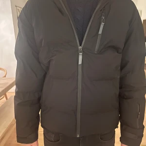Svart pufferjacka Jack & Jones Core - Svart pufferjacka från Jack & Jones Core med huva och flera dragkedjeförsedda fickor. Jackan har en clean look, lång ärm och normal passform. Perfekt för kalla dagar med sin tjocka och vadderade design. Snygg och enkel att matcha med jeans eller hoodies.
