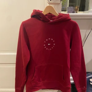 Vinröd hoodie från Tommy Hilfiger - Snygg röd hoodie från Tommy Hilfiger med logga framtill och klassisk känguruficka. Tröjan har huva och är tillverkad i ekologisk bomull. Perfekt för dig som gillar en stilren och bekväm look.