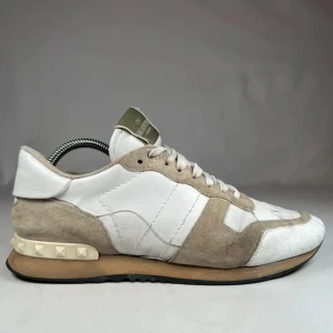 Valentino Garavani Rockrunner sneakers - Snygga Valentino Garavani Rockrunner sneakers i vitt och beige med coola nitar på hälen. Skorna har en mix av mocka och skinn, klassisk snörning och svart sula. Perfekta för dig som gillar exklusiva och trendiga sneakers med unik design. Är dock lite creazade.