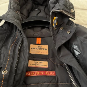 Svart Parajumpers pufferjacka med huva - Svart pufferjacka från Parajumpers med huva, orange detaljer och grov dragkedja. Jackan har flera fickor med lock och tryckknappar, samt quiltad insida. Snygg broderad etikett med 'Masterpiece Series' på insidan. Perfekt för kalla dagar.