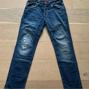 jeans från Jack & Jones W28 L32 - Säljer ett par blå jeans från Jack & Jones med slitningar och hål på benen. Klassisk femficksmodell med snygga detaljer på bakfickorna och normal passform. Jeansen har dragkedjegylf och är i ett mjukt denimtyg.