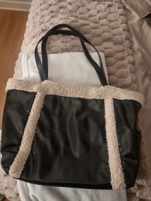 Svart stor väska med teddy-detaljer - svart lite större väska med mjuka teddy-detaljer i beige längs kanterna. Väskan har svarta handtag i syntetmaterial och dragkedja upptill. Insidan är rymlig med flera fack för småsaker. Perfekt för dig som vill ha praktisk väska.