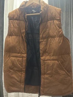 Brun dunväst från Color State - Säljer en brun dunväst från Color State i storlek M. Västen har en hög krage, två stora fickor framtill och stängs med dragkedja. Perfekt att slänga över en hoodie för extra värme. Enkel och stilren design.