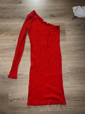 Röd oneshoulder klänning från NA-KD - Säljer en snygg röd ribbad klänning från NA-KD i storlek S. Klänningen har oneshoulder-design och lång ärm på ena sidan, vilket ger en cool och modern look. Materialet är mjukt och stretchigt.