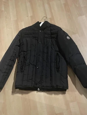 Svart quiltad dunjacka från Moncler - Svart dunjacka från Moncler med quiltad design och lång ärm. Jackan har ribbad krage, knappstängning framtill och två fickor med dragkedja. Märkespatch på ena ärmen och innerfoder med etikett och serietidningsmönster. Jackan kommer inte till användning och därför säljer jag den och den är perfekt för kalla dagar. Jackan är förmodligen äkta 