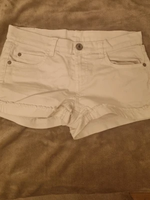 Vita shorts från KappAhl, storlek 158 - Säljer ett par vita shorts från KappAhl i storlek 158. De har klassisk femficksdesign, knapp och dragkedja framtill samt bälteshällor. Materialet är mjuk bomull och shortsen har en snygg, lite fransad kant nedtill. Perfekta för varma dagar!