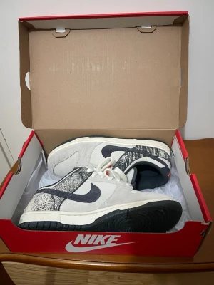 Nike Dunk Low med svart och vit splash - Nike Dunk Low sneakers i vit och grå canvas med svarta detaljer och splash-mönster på hälen. Swoosh-logga i svart, röd Nike-text på hälen och plös. Skorna har snörning och platt sula, samt lågt skaft. Perfekt för dig som gillar streetwear och unika detaljer. Säljer för att de var för stora och har bara haft på mig 1 gång 