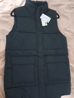 Svart dunväst från 157 - Svart dunväst från 157 med hög krage och två stora fickor framtill. Västen har quiltad design och stängs med dragkedja och tryckknappar. Perfekt för lager-på-lager och chill stil. Materialet är polyester och västen har en loose passform.