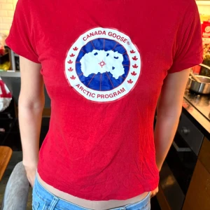 Röd Canada Goose t-shirt XS - Säljer en röd t-shirt från Canada Goose i storlek XS. T-shirten har ett stort tryck med logga och karta på bröstet, rund hals och korta ärmar. Materialet är mjuk bomull som känns skönt mot huden. Perfekt för dig som gillar streetwear och vill sticka ut.
