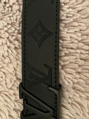 Svart IIvv bälte - Säljer ett svart bälte med präglade logga och monogramdetaljer längs hela bandet. Bältet har ett mattsvart  llvvv spänne och exklusiv känsla. Perfekt accessoar för att lyfta vilken outfit som helst.