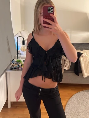 Svart volangblus från Amisu - Säljer en svart blus från Amisu i storlek M. Toppen har tunna axelband, volanger och knytning framtill. Den är croppad och har en luftig, flowy känsla som gör den perfekt till jeans eller kjol. Använd 2 gånger 