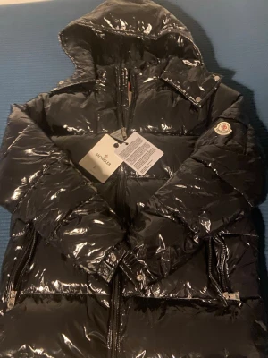 Moncler maya - Svart pufferjacka från Moncler med glansig finish, huva och dragkedjor framtill. Jackan har Moncler-logga på ärmen och flera snygga detaljer som tryckknappar och fickor. Perfekt för dig som vill ha en stilren och varm jacka med exklusiv känsla. Finns bilder ich QR kod NFT tag.