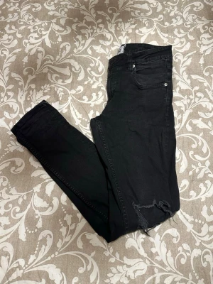 Svarta skinny jeans från Lager 157, M - Svarta jeans från Lager 157 i modellen Rocket, storlek M. Skinny fit med slitna detaljer och hål vid knäna för en edgy look. Klassisk femficksdesign och knappstängning. Materialet är jeans med stretch för skön passform.