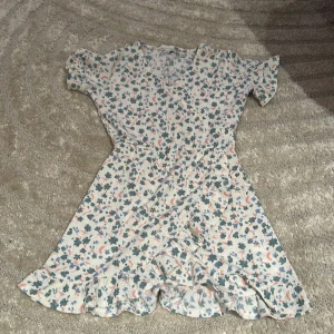 Blommig kortklänning från lager 157 - Supersöt kortklänning från Zara med blommigt mönster i grönt, rosa och blått på ljus botten. Klänningen har v-ringning, korta ärmar med lätt puff och volangkant nertill. Perfekt för sommarens alla dagar!