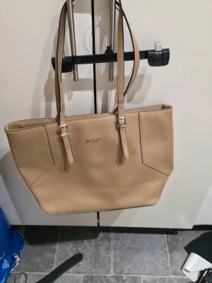 Beige handväska från Guess - Snygg beige handväska från Guess med stilrena silverdetaljer och justerbara axelremmar. Väskan är tillverkad i skinnimitation med en strukturerad yta och har en klassisk, fyrkantig form. Perfekt för dig som gillar enkel och tidlös design.