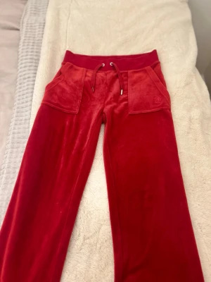 Röda byxor från Juicy Couture😽 - Säljer ett par röda mjukisbyxor från Juicy Couture med snörning vid midjan. Knappar & silverfärgade detaljer. Materialet är mjukt och glansig som känns lyxigt.    Viktigt att tillägga att det är upplagda där nere cirka 3 cm och tappat en grej som ni kan se på sista delen men ser fortfarande as snygga ut💕