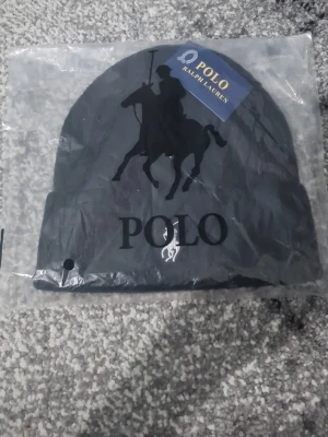 Svart ribbad mössa från Polo Ralph Lauren - Snygg svart mössa från Polo Ralph Lauren med ribbad struktur och klassisk broderad logga framtill. Perfekt för att hålla värmen och samtidigt se stilren ut. Mössan har uppvikt kant och är tillverkad i mjukt material.