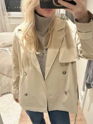 Beige trenchcoat från H&M med bälte - Stilren beige trenchcoat från H&M med dubbelknäppning, bred krage och bälte i midjan. Jackan har långa ärmar med spänndetaljer vid handlederna och klassiska detaljer som axelklaff och slät finish. Perfekt för dig som gillar en tidlös look.