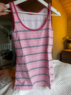 Randig rosa linnetopp - Säljer en fräsch rosa linnetopp med tunna axelband och rund halsringning. Toppen har ett randigt mönster i rosa, vitt och mörkblått, med kontrastkant i rosa runt hals och ärmhål. Perfekt för varma dagar och enkel att matcha med jeans eller shorts.
