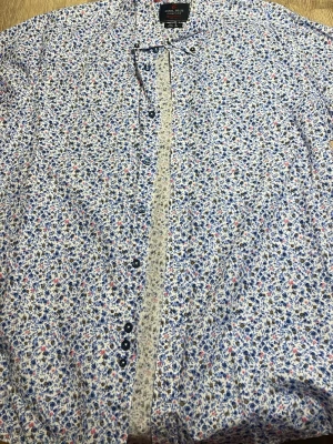 Blommig skjorta från Cool Blue Company - Säljer en blommig skjorta från Cool Blue Company i storlek L. Skjortan har ett mönster med små blå, rosa och bruna blommor på vit botten, knappar framtill och är tillverkad i 100% bomull. Regular fit för en skön och avslappnad passform.