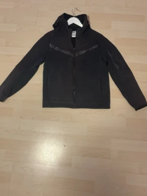 Svart Nike tech från Nike XL(158-170cm) - Svart Nike tech/hoodiejacka i storlek XL(barn)med hel dragkedja framtill och diskreta svarta detaljer över bröstet. Jackan har lång ärm, huva och är gjord i mjukt bomullsmaterial. Perfekt för chill eller träning, enkel och stilren design.