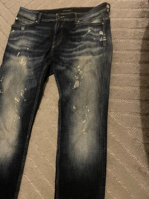 Mörkblå jeans från SKOG/DENIM 33/34 - Säljer ett par mörkblå jeans från SKOG/DENIM i storlek 33/34. Jeansen har slitna detaljer och ljusa partier framtill för en riktigt cool, använd look. Klassisk femficksmodell med knapp och dragkedja. Perfekta för dig som gillar streetwear-vibe.