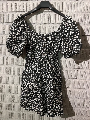 Svart blommig shortsdress från H&M - Supersöt svart shortsdress från H&M med vita blommor och puffiga kortärmar i mycket fint skick! Klänningen har knappar framtill och en dragkedja och även fickor! Jättefin att ha vid festtillfällen!