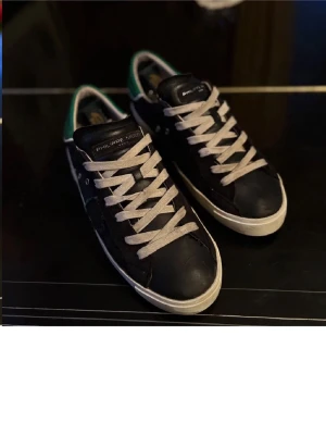 Philippe Model svarta sneakers med grönt - Svarta sneakers från Philippe Model med gröna detaljer på hälen och vita snören. Skorna har en rund tå, platt sula och en skön innersida. Materialet är skinn och de har en klassisk sneakerlook/grishlook med märkets logga på plösen. 