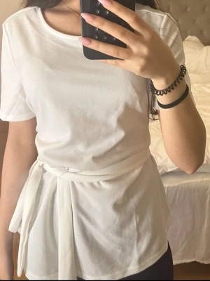 Vit omlottopp med knyt från SHEIN - Säljer en vit omlottopp från SHEIN med rund hals och korta ärmar. Toppen har ett brett knytband i midjan som ger en snygg siluett. Storlek M men funkar även som S och XS för en pösigare look som på bilden.