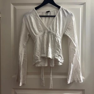 Vit omlottopp med knytband - Säljer en vit omlottopp med långa ärmar och v-ringning. Toppen har knytband i midjan som ger en snygg siluett och en lätt volangkant nedtill. Perfekt till jeans eller kjol för en fräsch look. Från gina tricot, storlek S